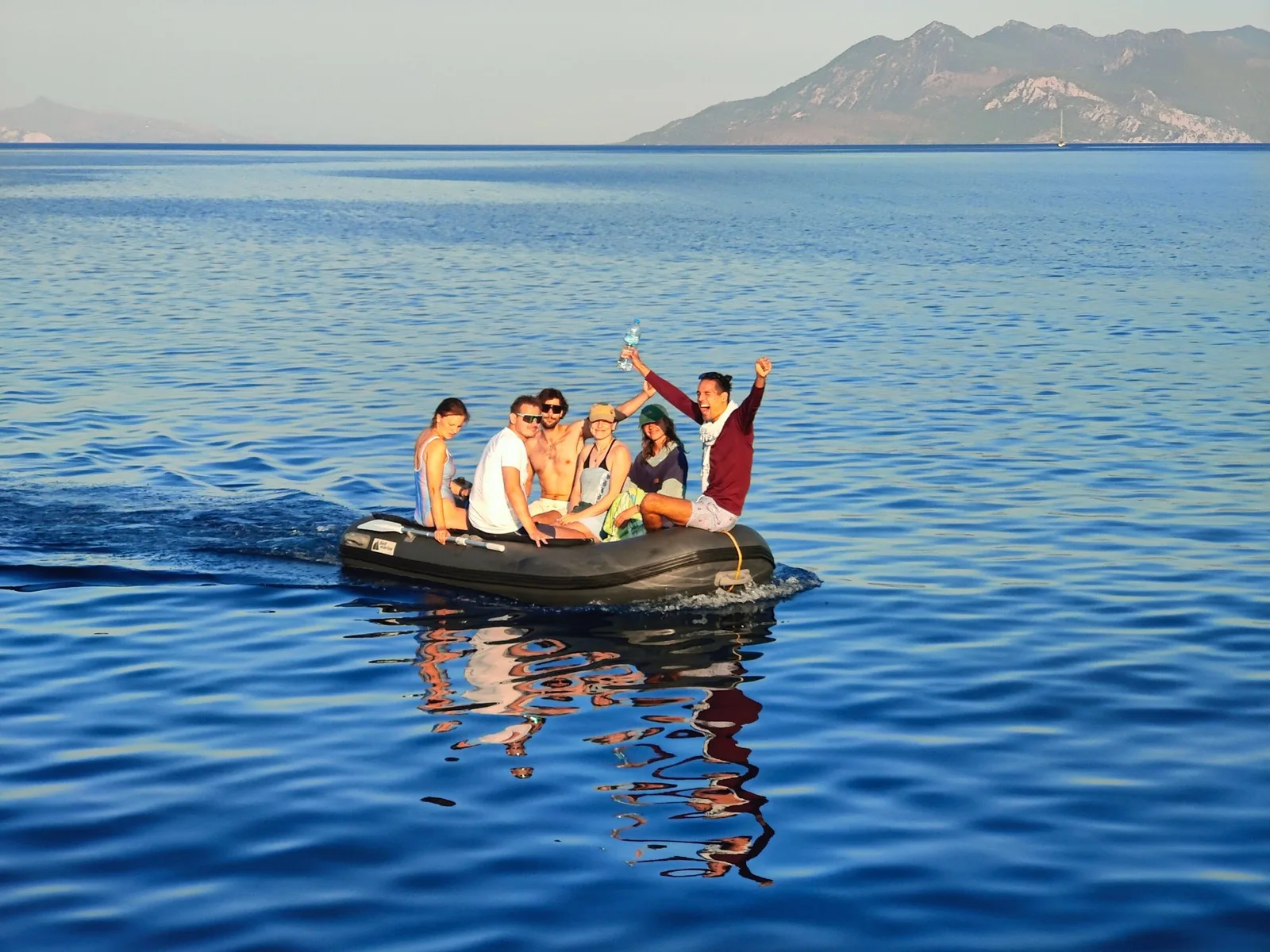 Freunde jubeln in einem Beiboot auf ruhigem, blauem Wasser vor einer griechischen Insel